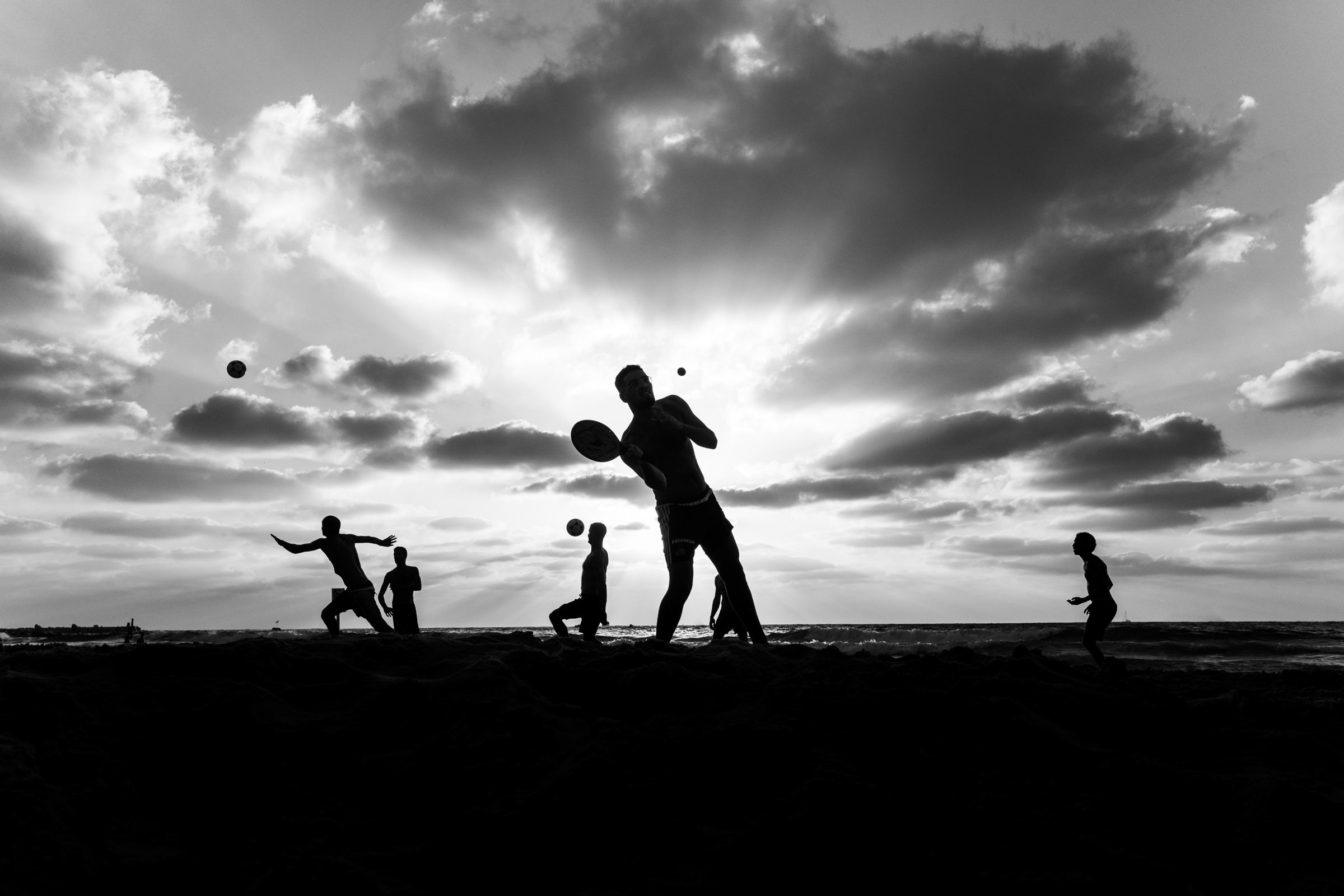 Black-and-white photograph: Silhouettes of people playing with rackets and balls on a beach at sunset
