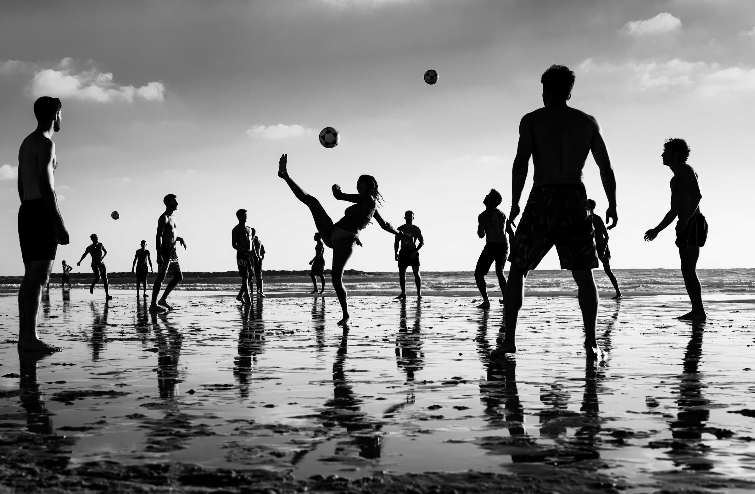 Black-and-white photograph: Silhouetted people play with balls on a wet beach at sunset