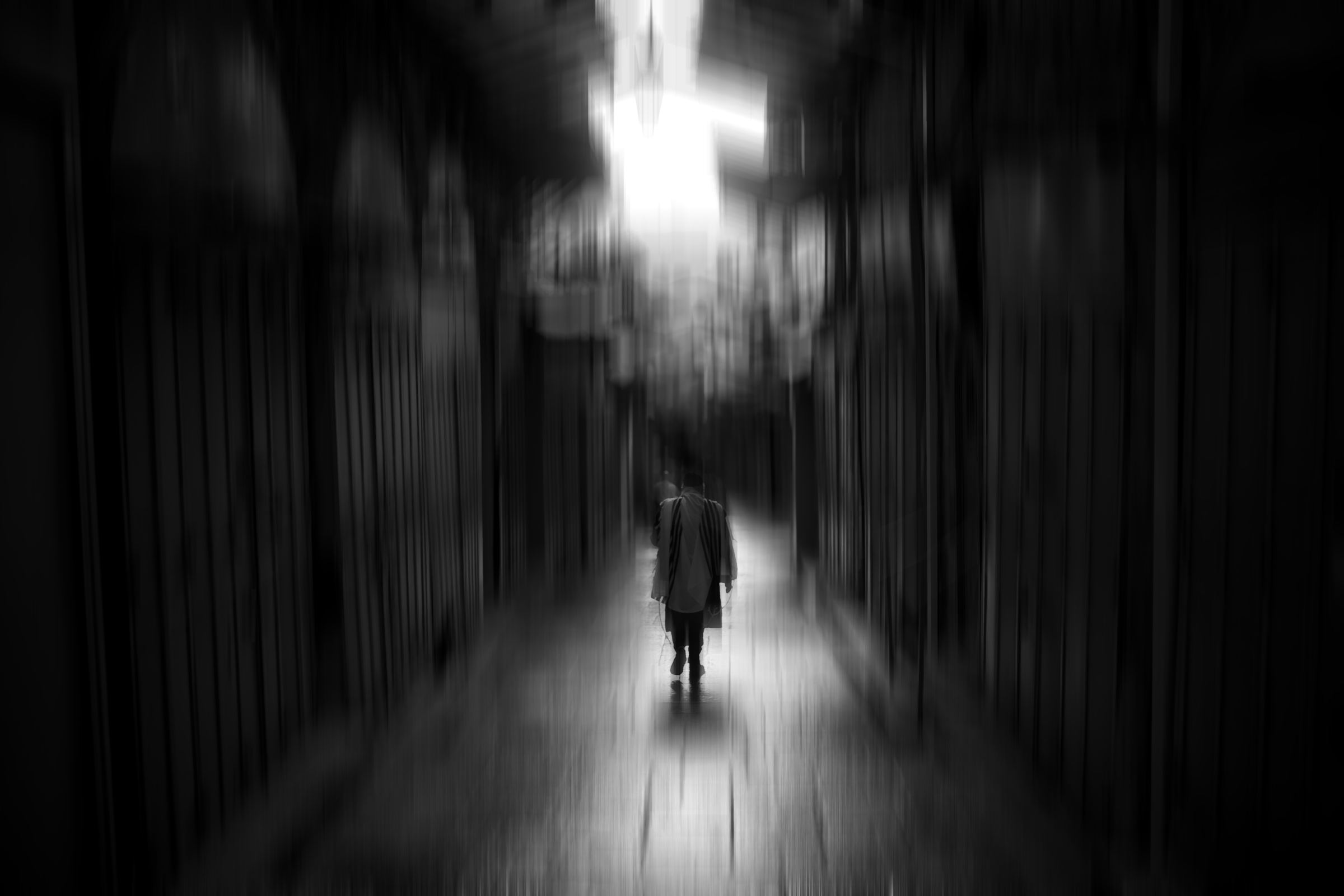 Black-and-white photograph: A figure in a prayer shawl walks away down a dark, blurred alley toward a bright light