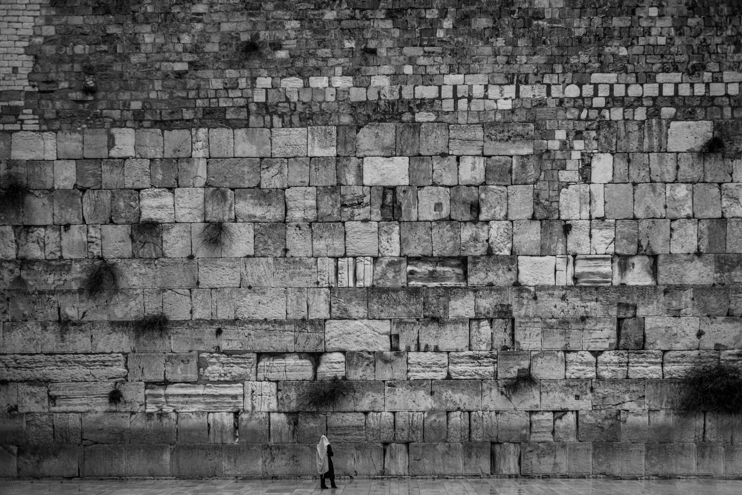 Black-and-white photograph: A lone figure in a prayer shawl stands at the foot of a vast wall of ancient stone blocks