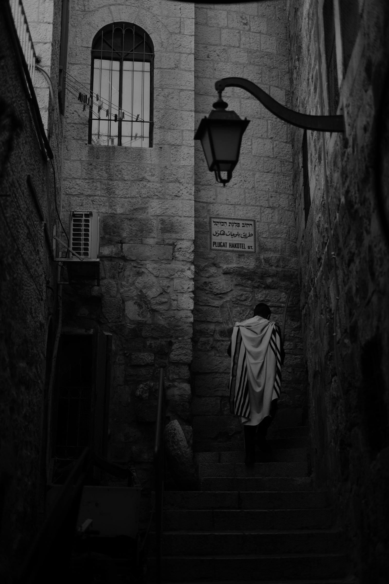 Black-and-white photograph: A man in a prayer shawl climbs stone steps in a narrow alley, beneath a street lamp and a sign reading Plugat Hakotel St.
