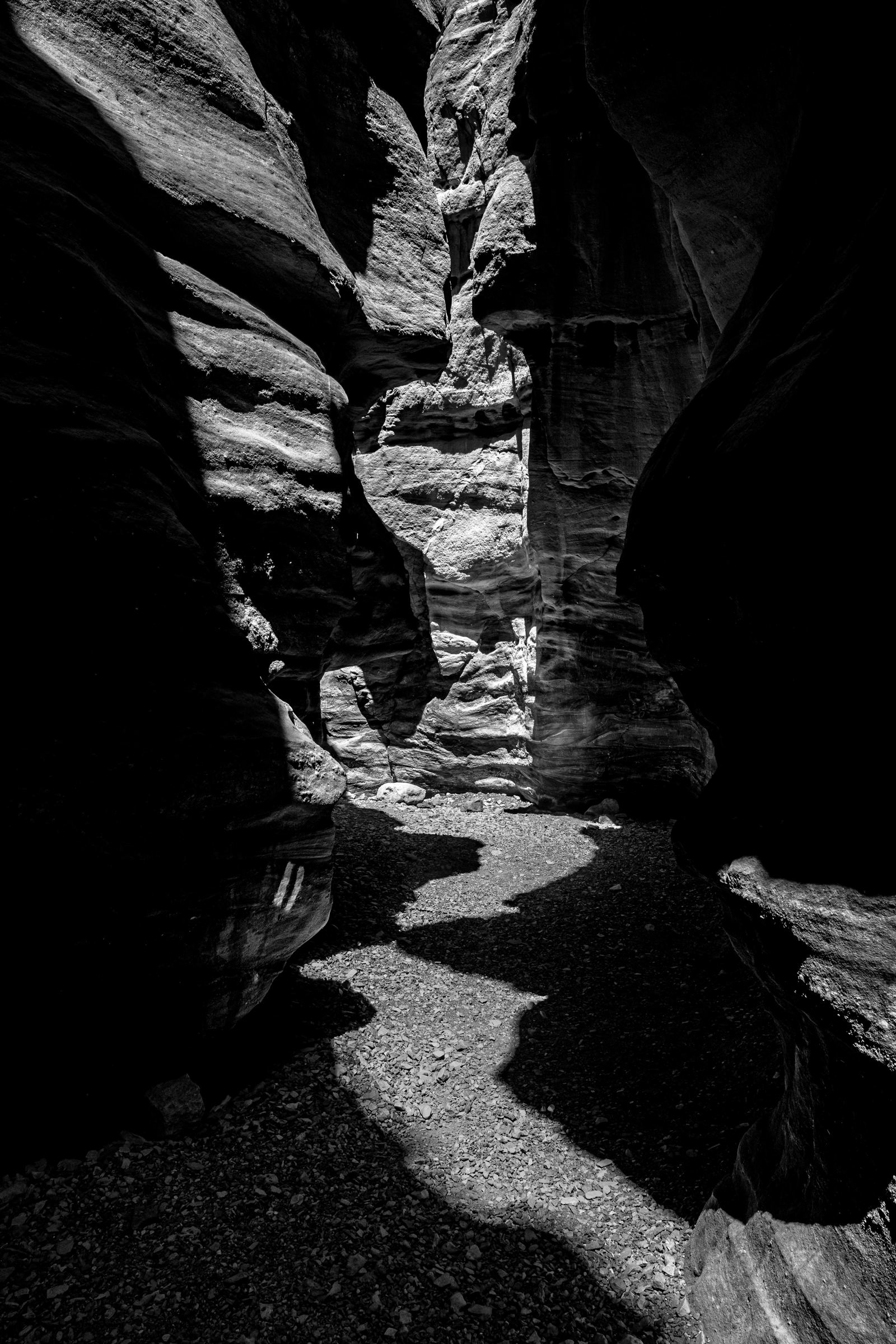 Black-and-white photograph: Light falls across the floor and walls of a narrow sandstone canyon