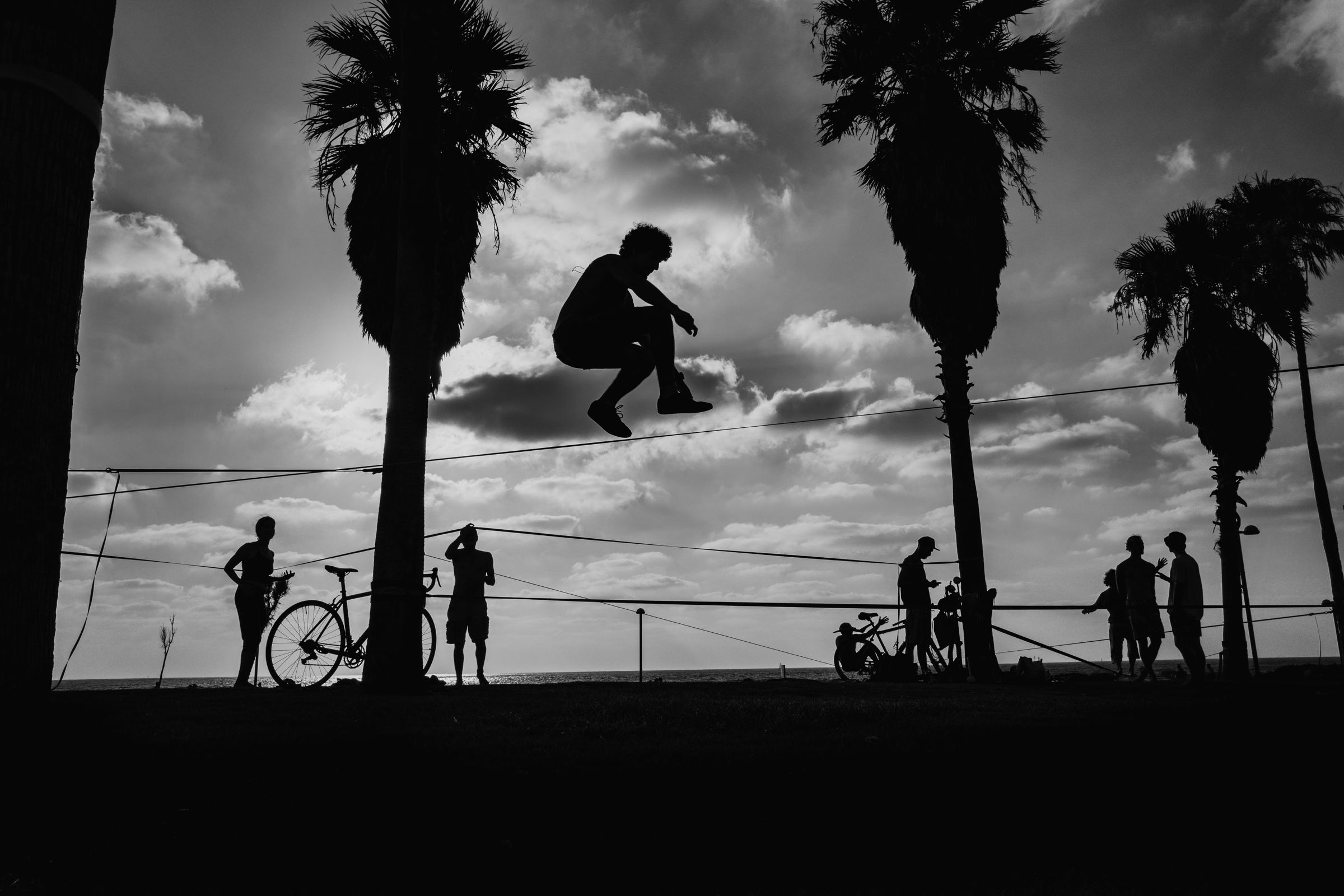 Black-and-white photograph: A man leaps on a slackline between palm trees at sunset, in silhouette, while people watch below