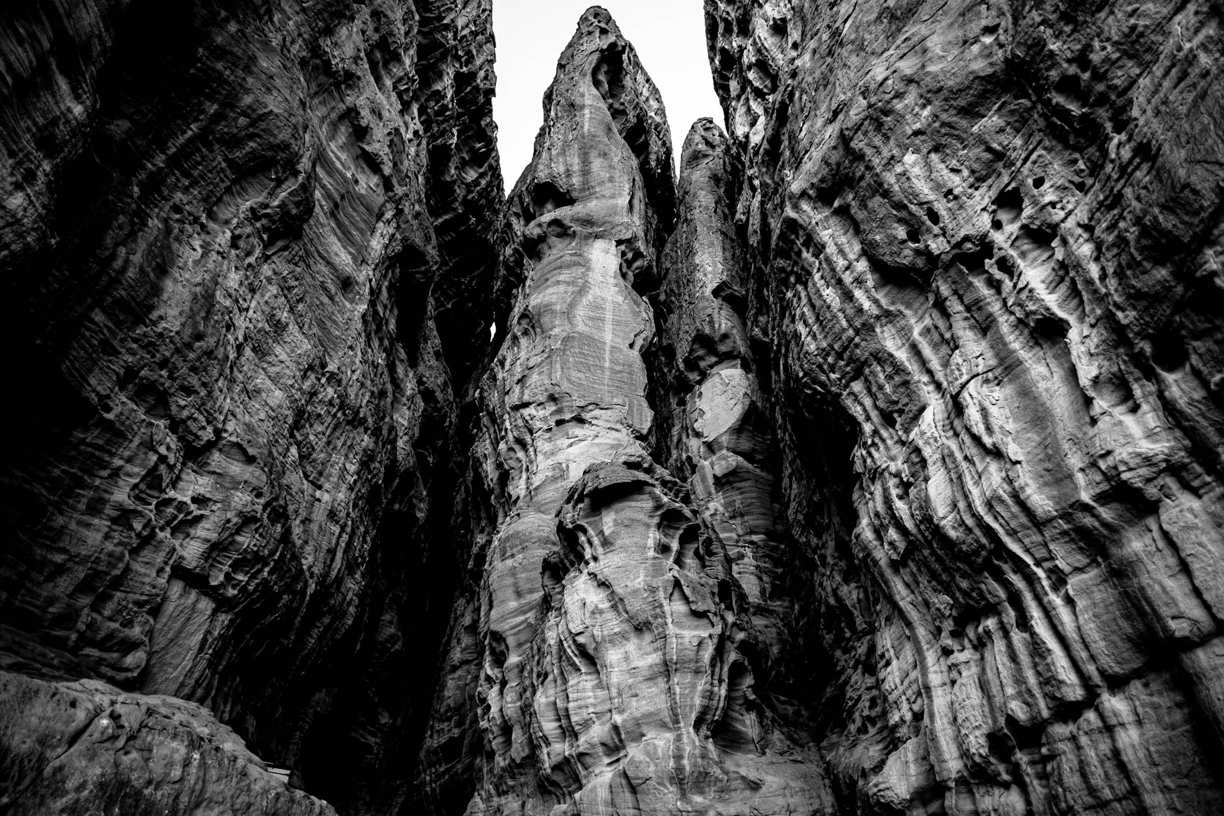 Black-and-white photograph: Looking up between two walls of sandstone at a tall rock column, with a narrow strip of sky above