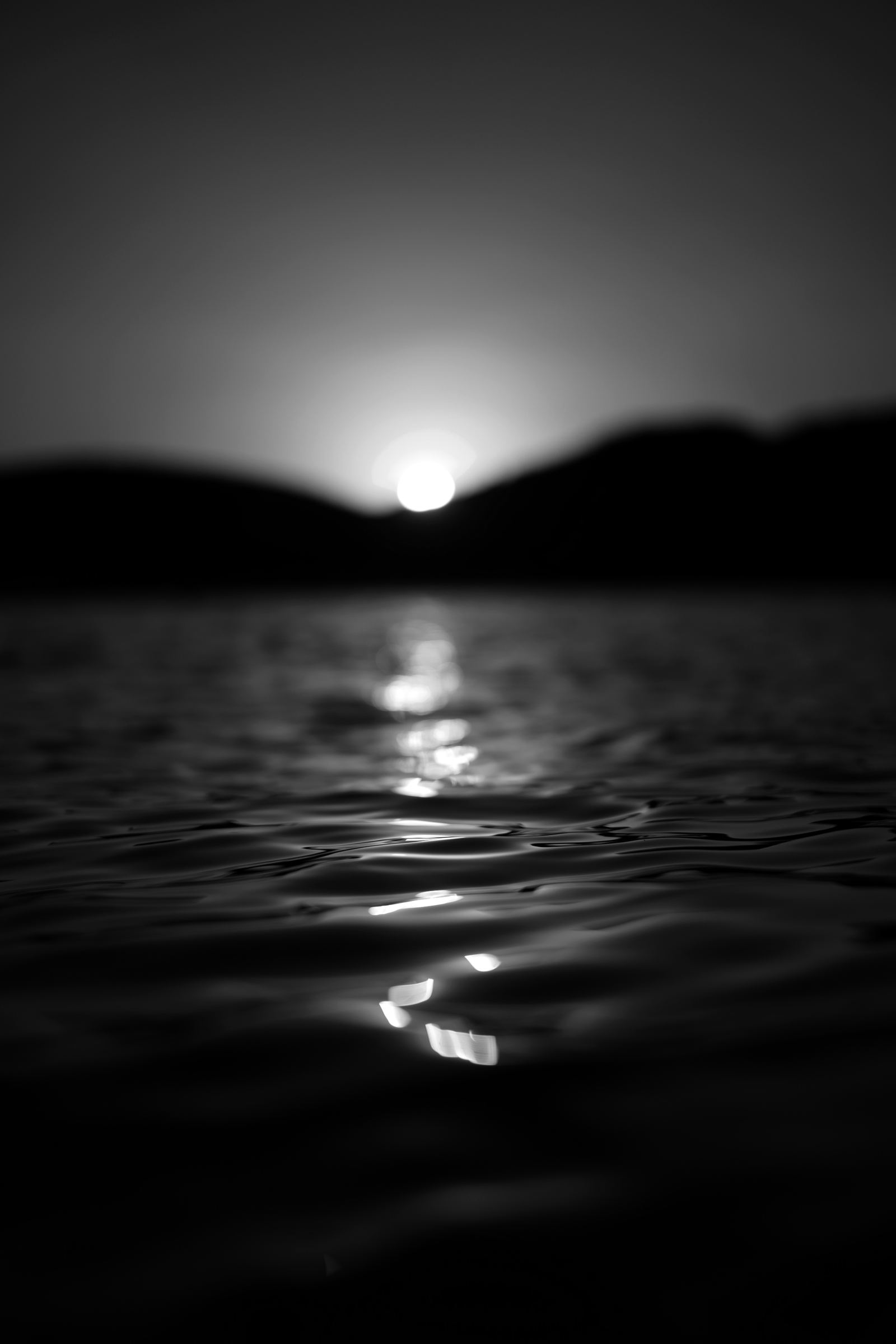 Black-and-white photograph: The sun just above a dark hill, its light reflected on rippling water