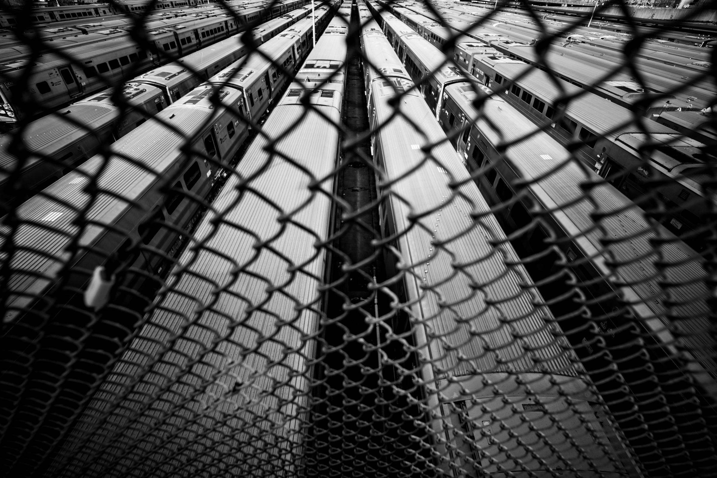 Black-and-white photograph: Rows of parked train cars seen from above through a chain-link fence